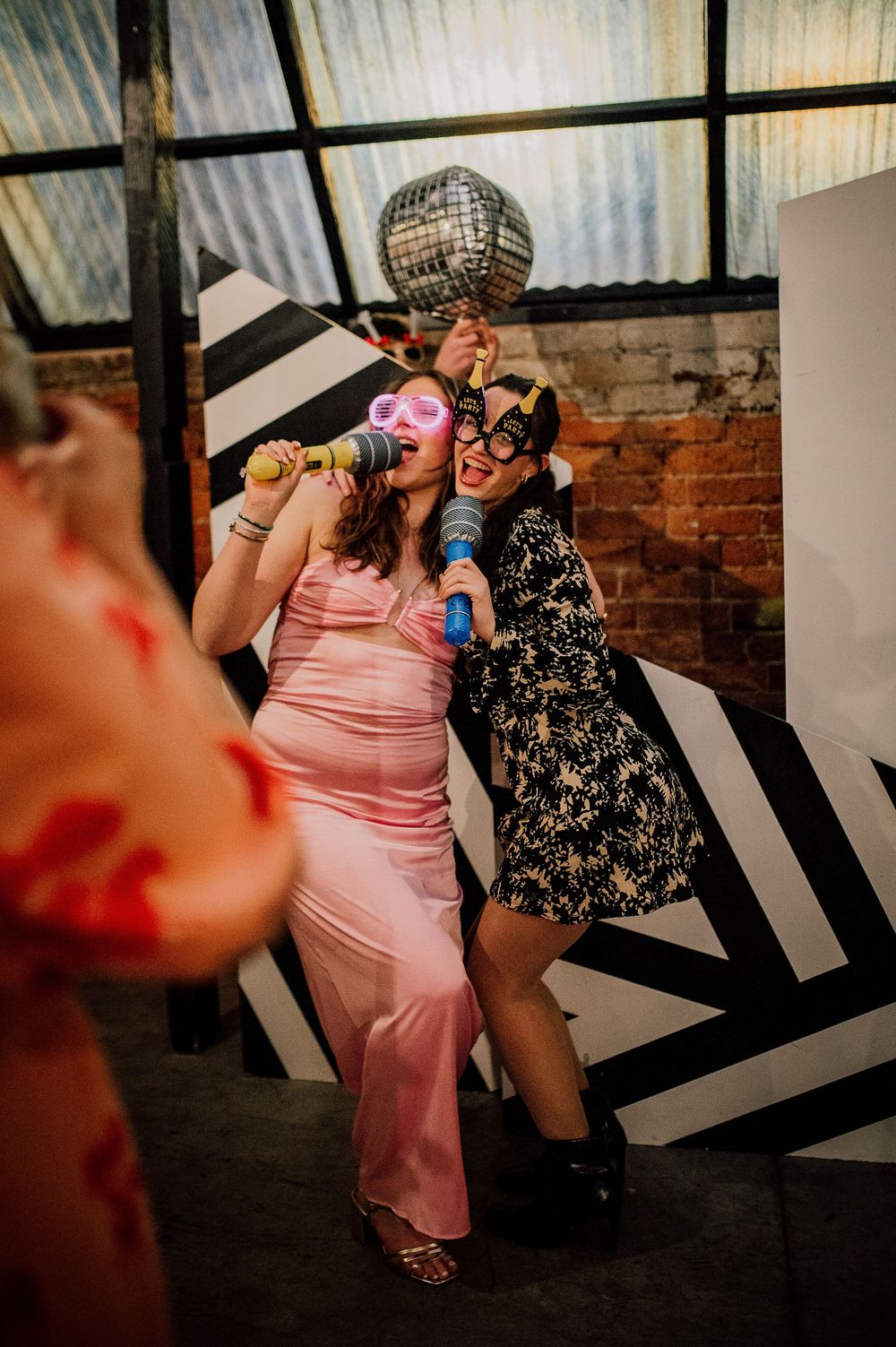 Two party guests dancing and posing under a disco ball at a celebration with exposed brick walls and geometric decor.