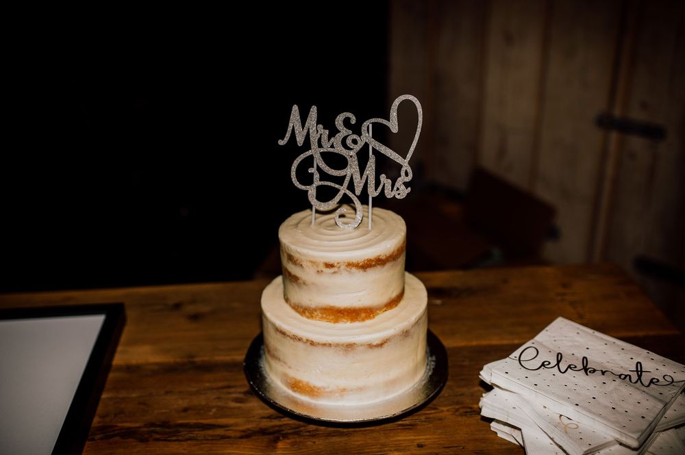 A rustic three-tier naked wedding cake with a Mr & Mrs topper sits on a wooden table beside a guest book.