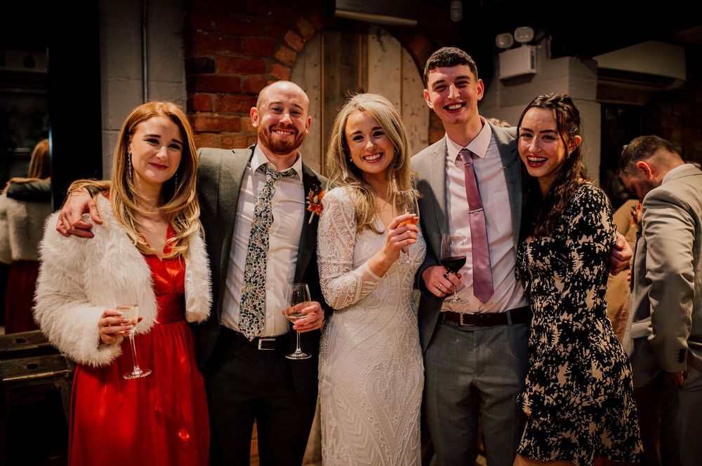 A group poses playfully for photos at a formal event with brick walls and festive attire.