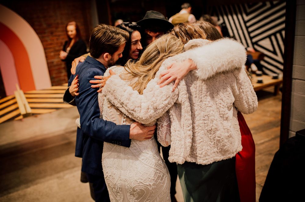 A group of people embrace in a warm hug at an indoor venue with brick walls and geometric patterns.