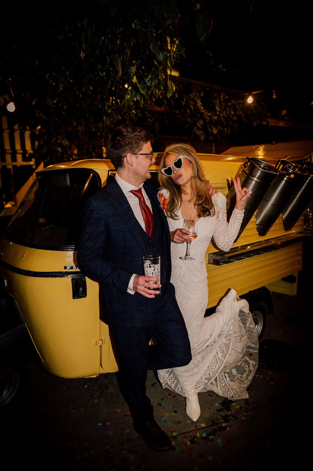 A couple in formal wedding attire poses playfully near a vintage yellow car during an evening celebration.