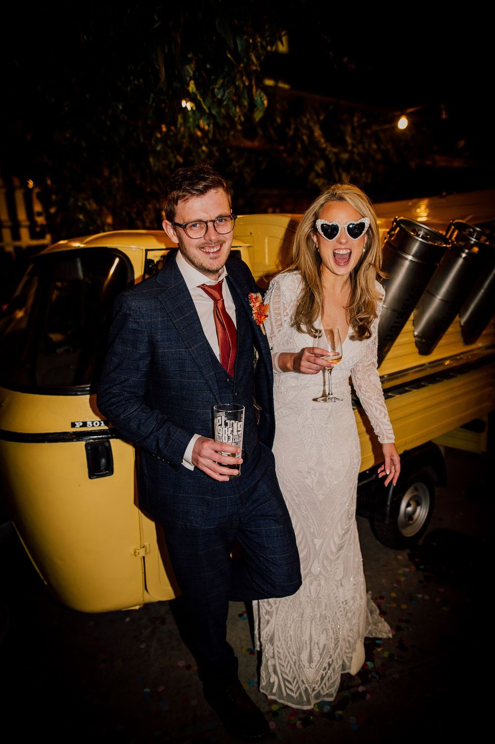 A couple in formal attire poses next to a vintage yellow car at night during a celebration.