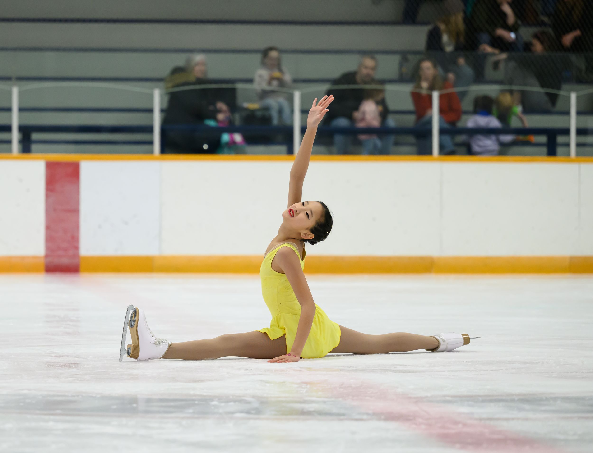 Figure skater in yellow dress performs graceful split pose on ice during competitive performance.