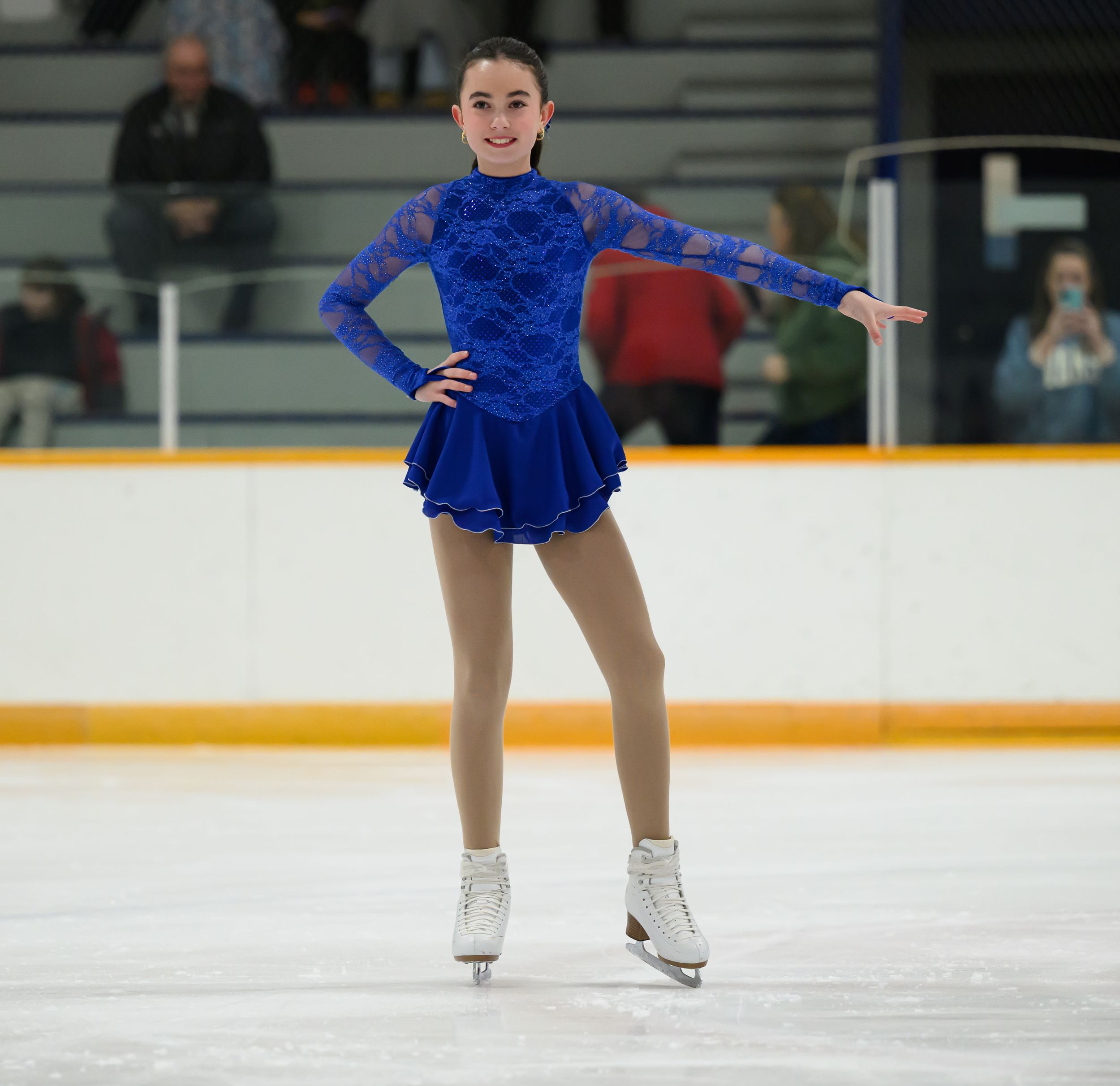 Figure skater in royal blue dress performs on ice rink while striking elegant pose during competition.