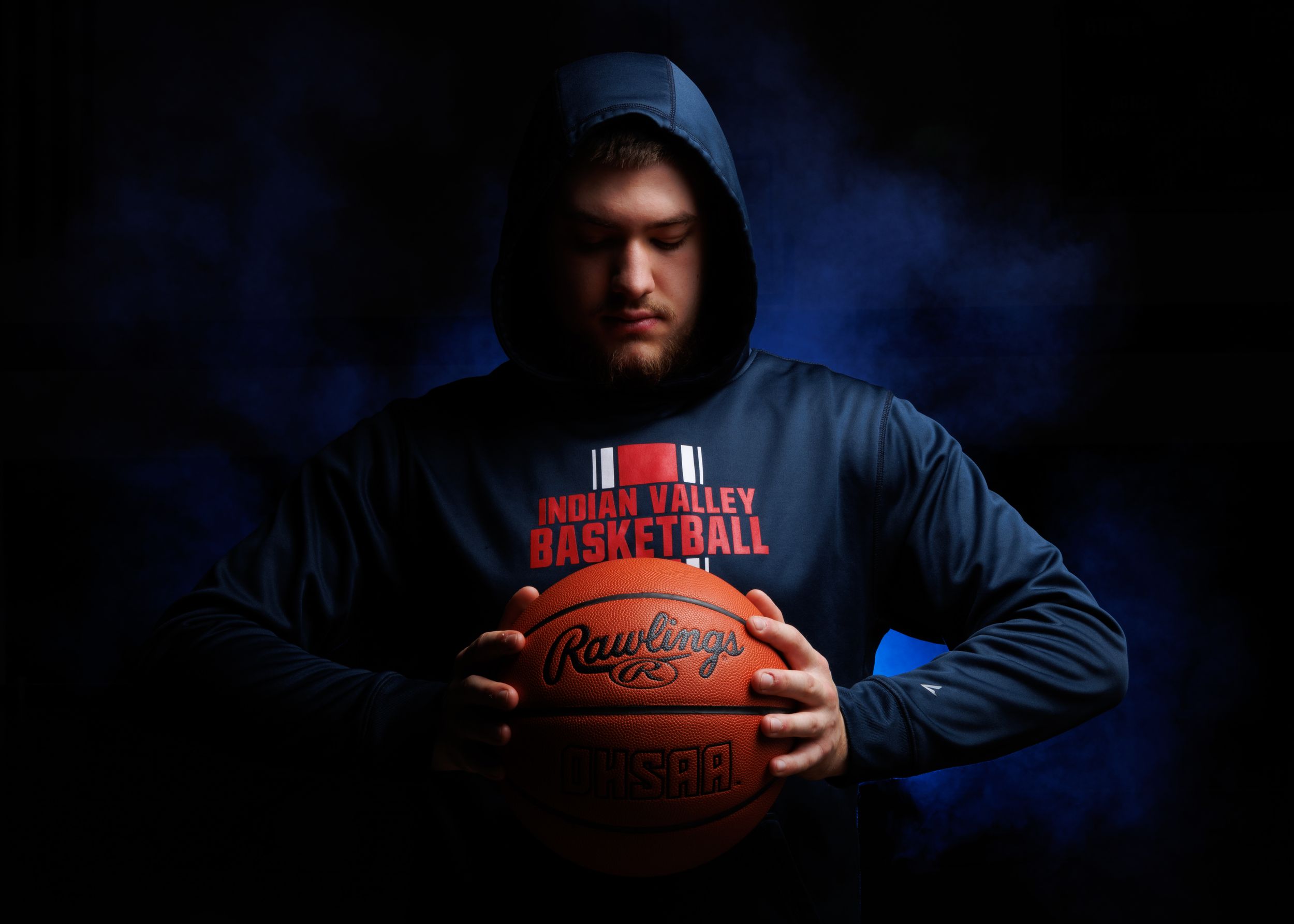 Basketball player in dark hoodie holds ball in moody blue lighting.