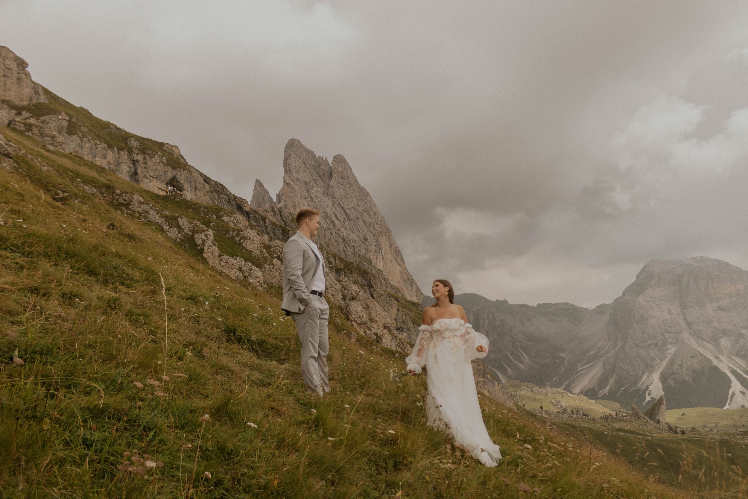 A couple in white attire stands on a grassy mountainside with dramatic Dolomite peaks and moody clouds in the background.