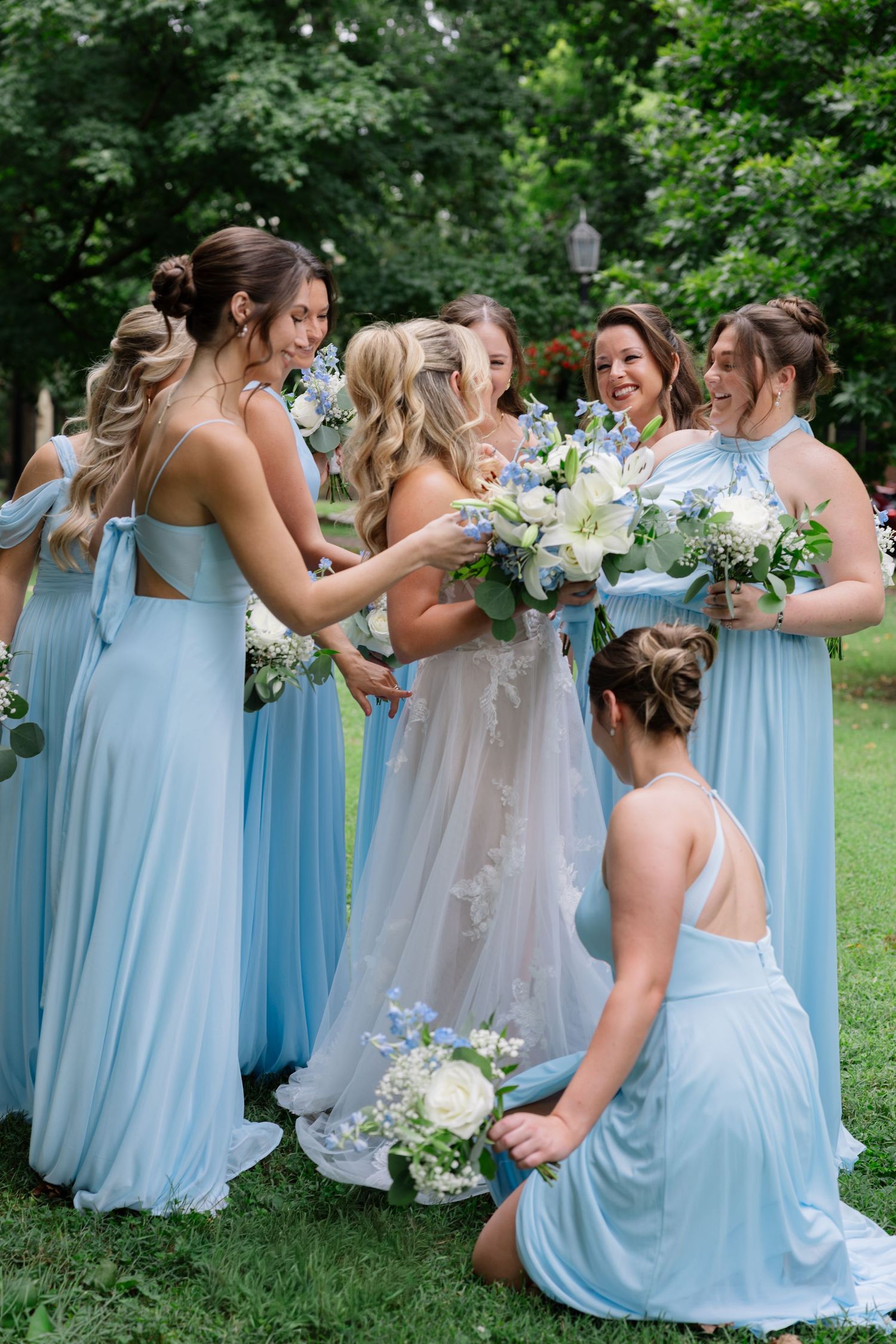 Bridesmaids in flowing light blue dresses gather outdoors for candid wedding photos in a garden setting.