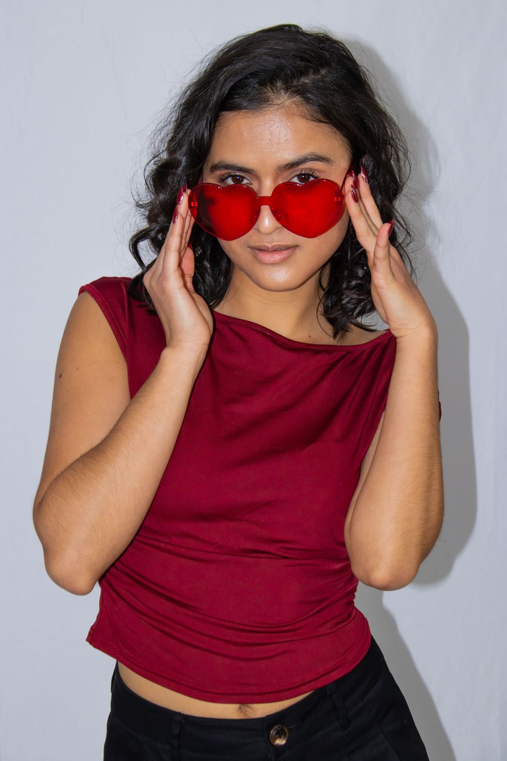 A person in a burgundy sleeveless top and black pants poses playfully with red sunglasses against a white background.