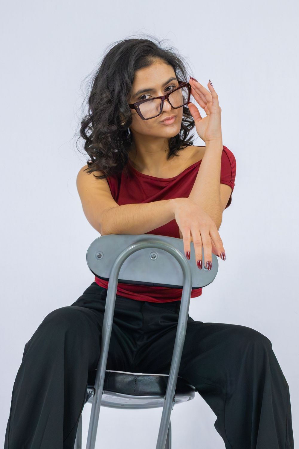 Stylish person in red top and black pants poses confidently on chair while adjusting glasses against white background.