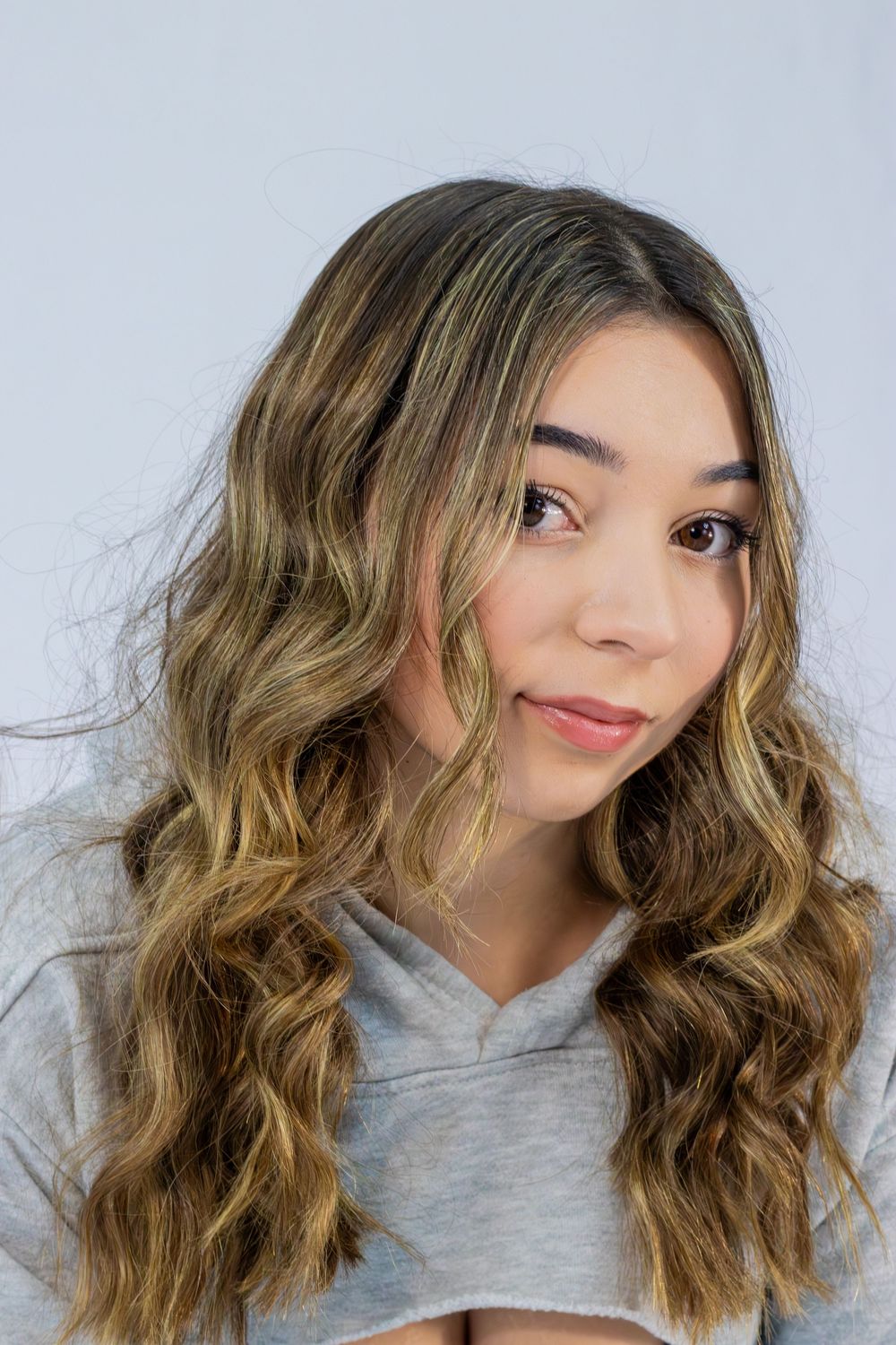 Model with wavy highlighted hair poses in gray top against minimalist white background.