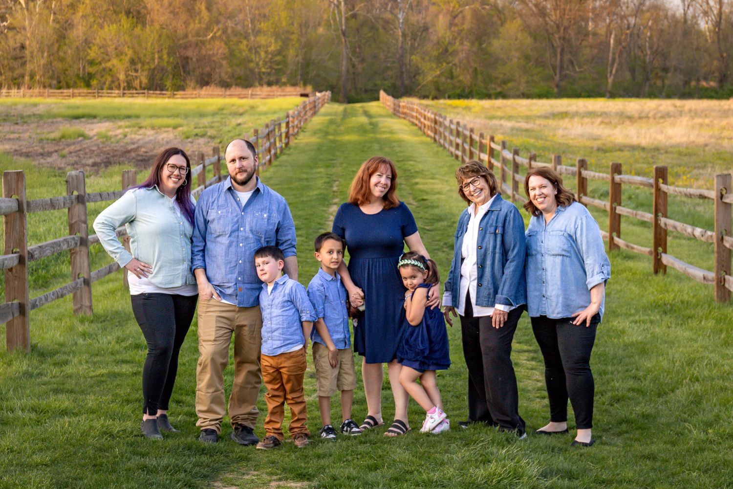 Large family standing together on a grassy field with wooden fences during autumn season.