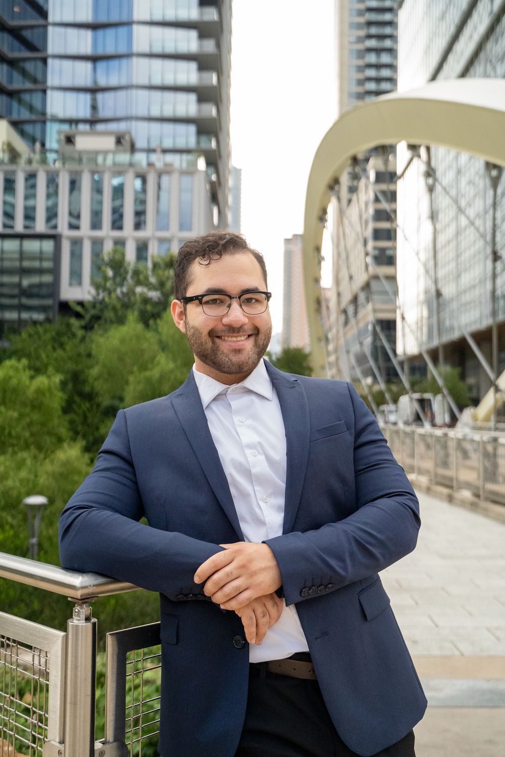 Professional in a navy suit and white shirt poses confidently on an urban rooftop terrace with city buildings in background.