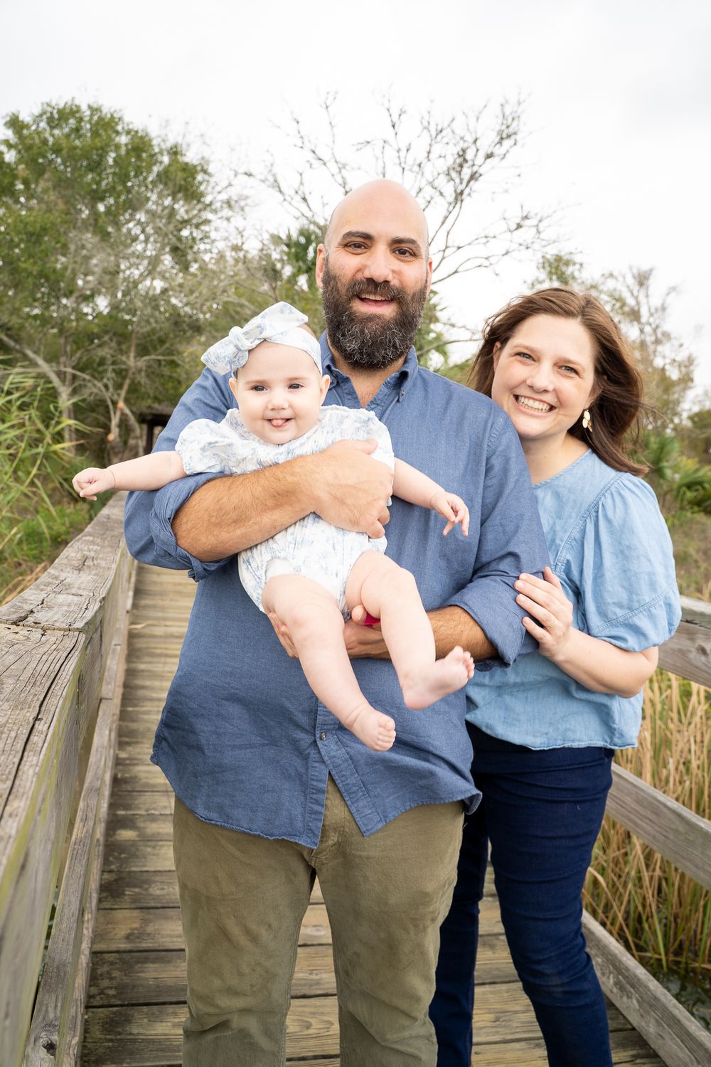 Happy family portrait on wooden bridge, bald bearded dad holding smiling baby girl, mom in blue top, outdoor nature setting.
