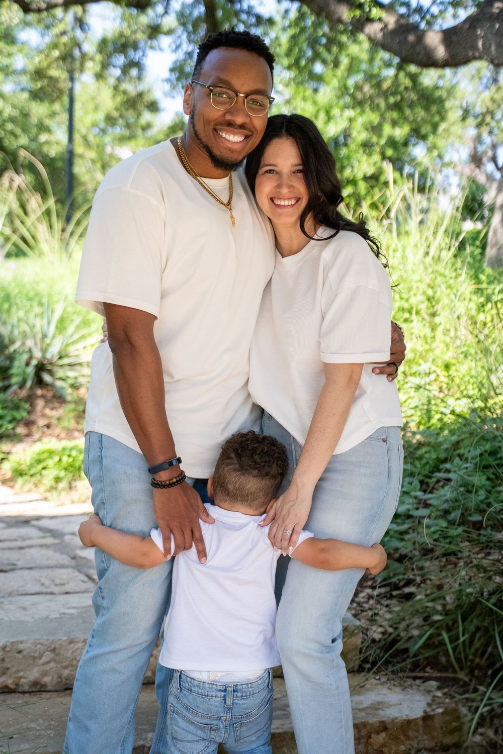 A casual family photoshoot in a sunny garden setting wearing matching white shirts and blue jeans.