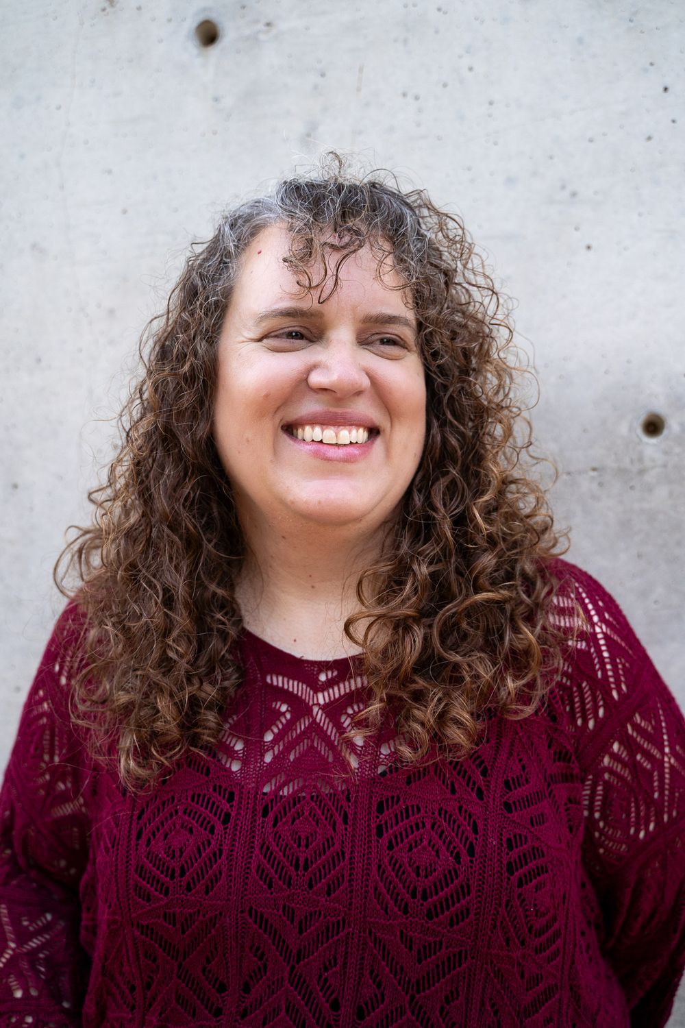 Smiling woman with curly brown hair wearing a red lace top, posing against a concrete wall.