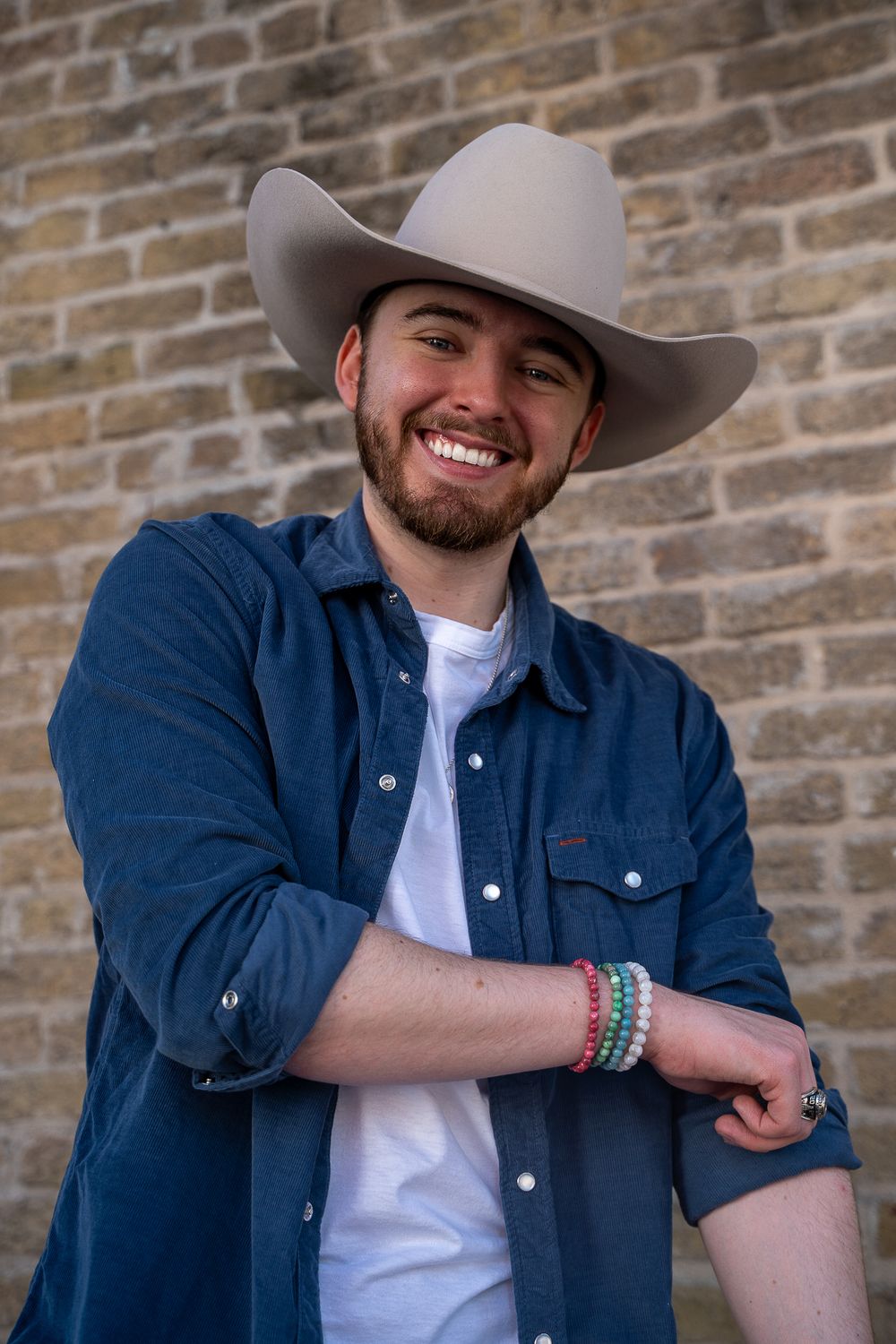 Smiling young man with beard wearing cowboy hat, blue denim shirt, and colorful bracelets against brick wall.
