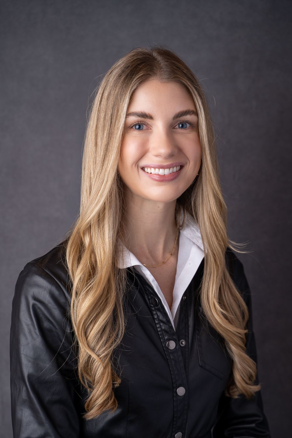 Young professional woman with long blonde hair smiling confidently in a black leather jacket and white shirt.