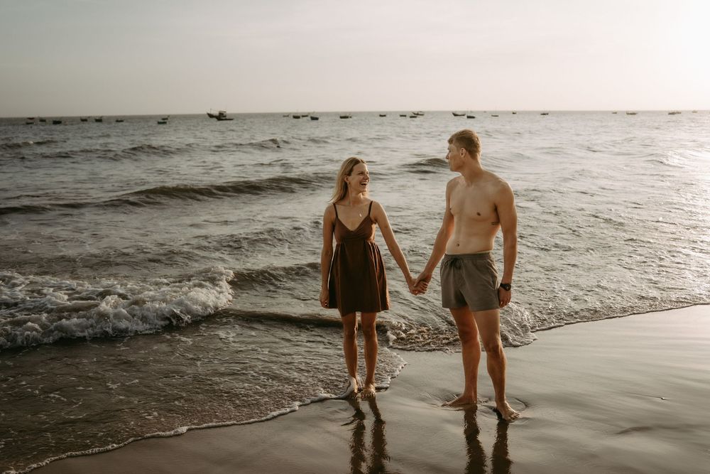 A couple holds hands while walking along the beach shoreline at sunset with waves rolling in.