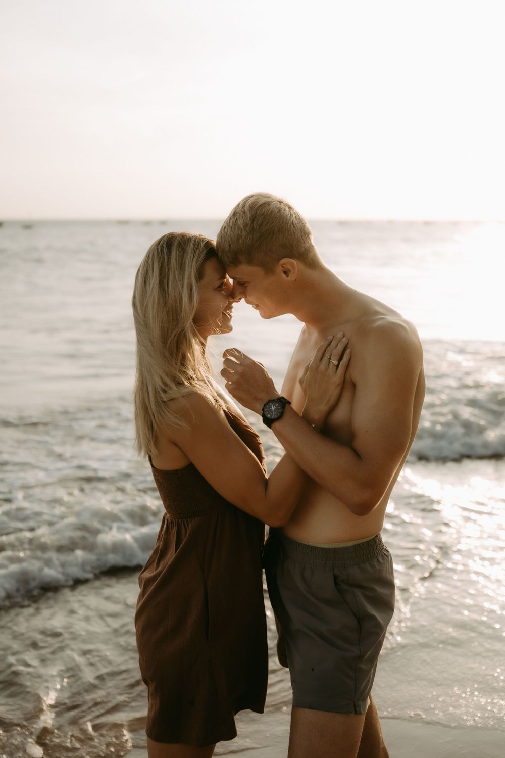 Romantic couple embracing on beach at sunset with waves crashing behind them.