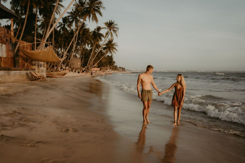 Tropical beach scene with palm trees lining the sandy shore as people walk along the water's edge at sunset.
