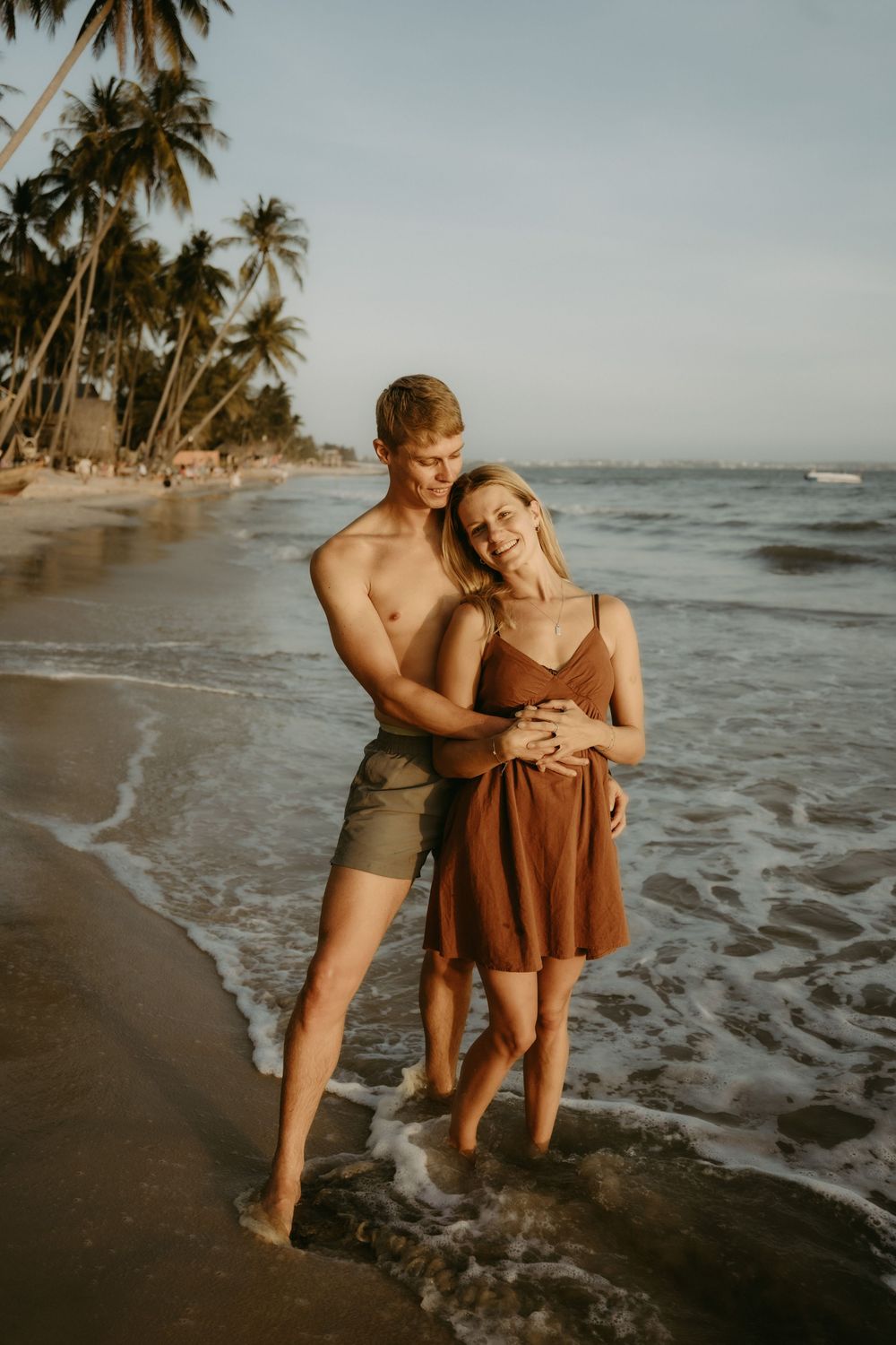 Couple embracing on a tropical beach with palm trees and ocean waves at sunset.