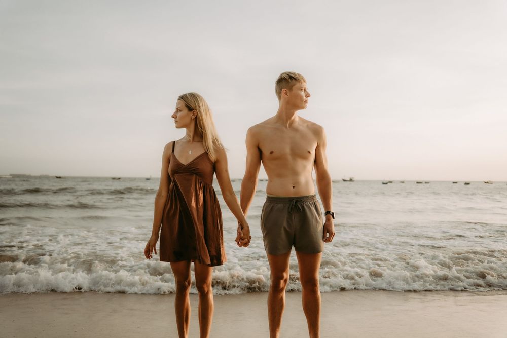 A couple walks hand in hand along a misty beach shoreline at sunset with waves crashing beside them.