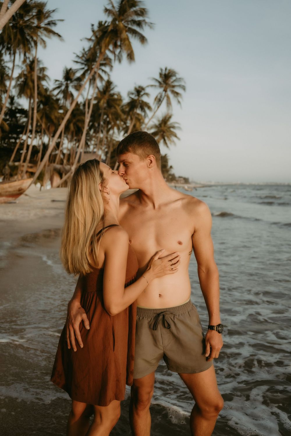 A couple shares an intimate moment on a tropical beach with palm trees swaying in the breeze at sunset.
