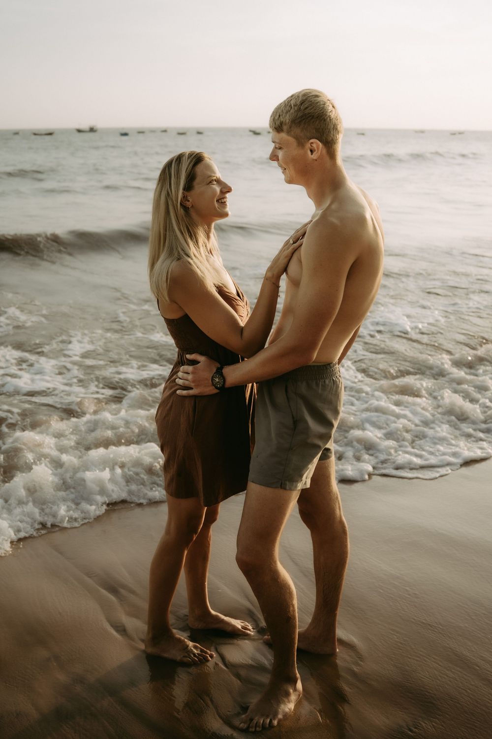 A romantic couple embraces on a sandy beach at sunset with ocean waves in the background.