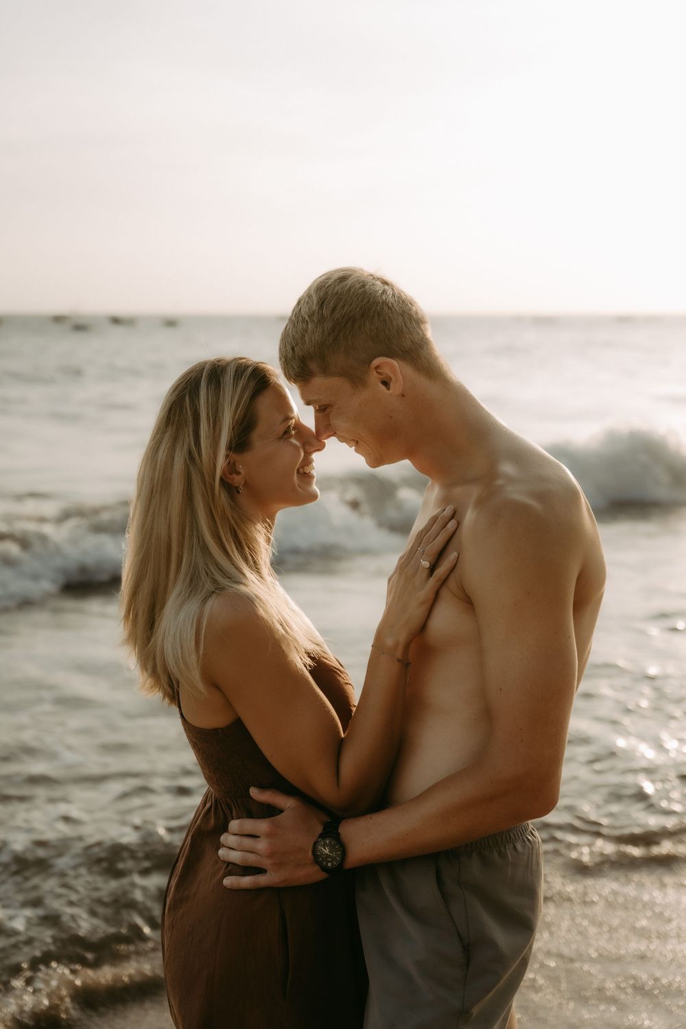 A romantic couple embraces on a beach at sunset with waves crashing behind them in a series of intimate poses.