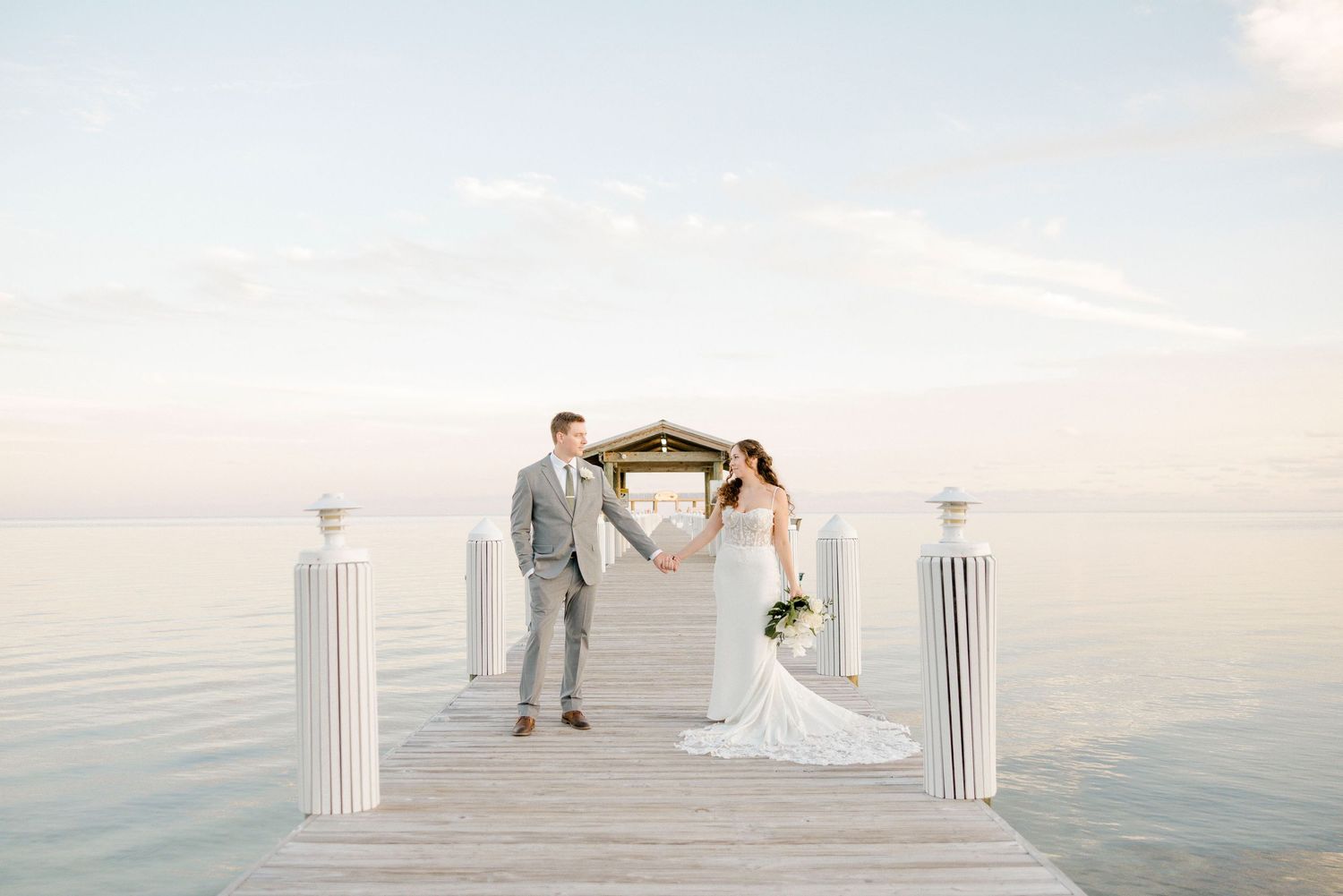 A couple in wedding attire walks along a serene beach at sunset with white pillars framing the romantic scene.