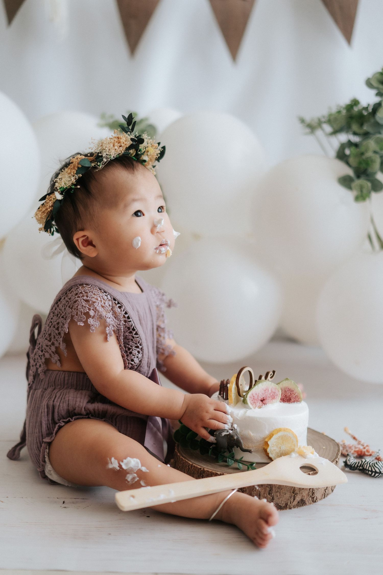 Baby in brown romper sits on white backdrop with cake during milestone photoshoot session.