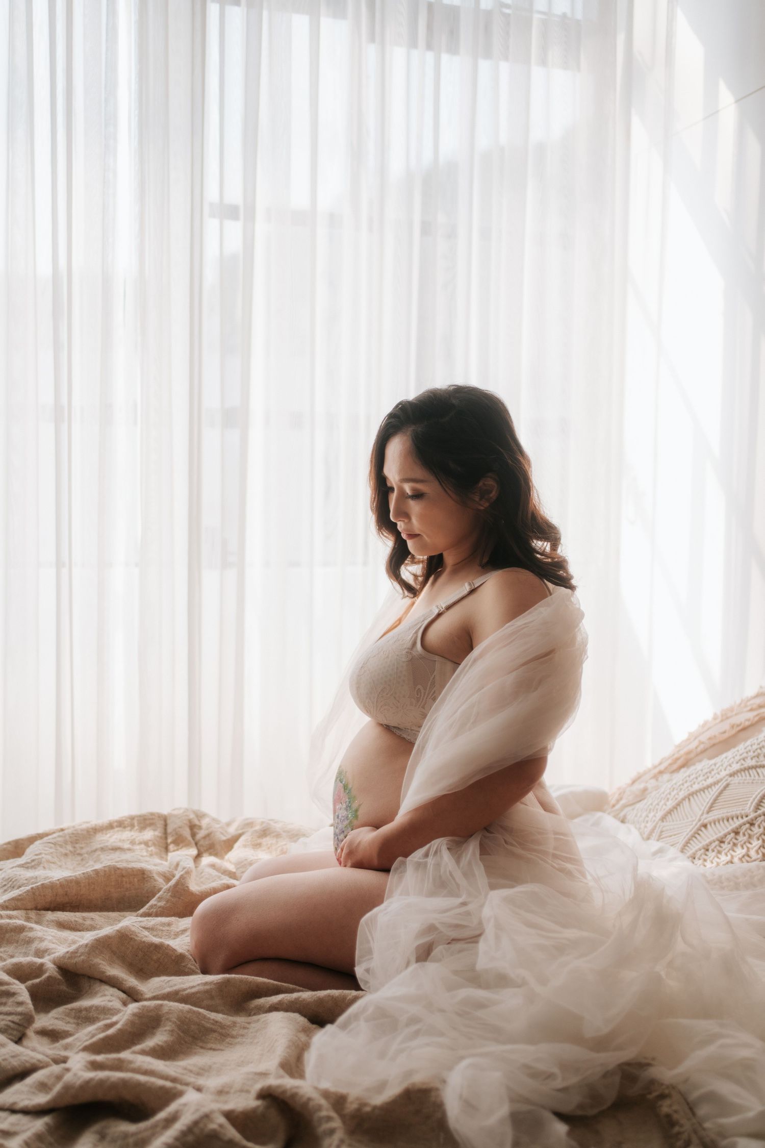 Ethereal maternity photo with soft white draping fabric and natural window light illuminating a peaceful bedroom scene.