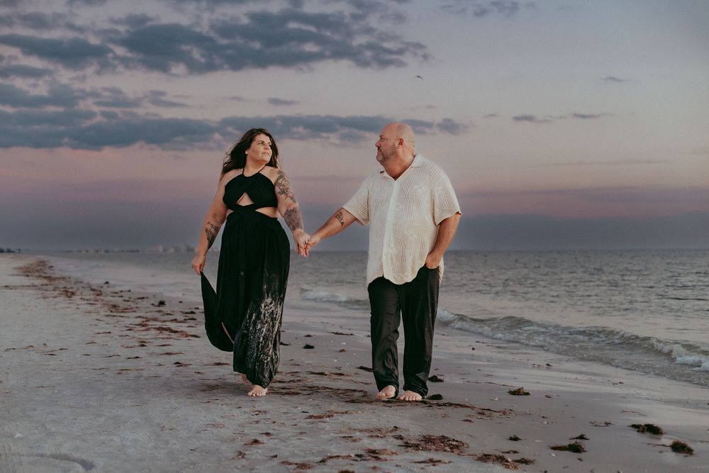 Two people walk hand in hand along a beach at sunset with soft pink and purple sky over the ocean.