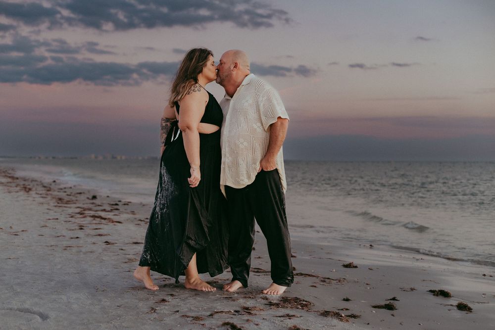 A couple sits together on rocks by the ocean during sunset, creating a romantic silhouette against the pink sky.