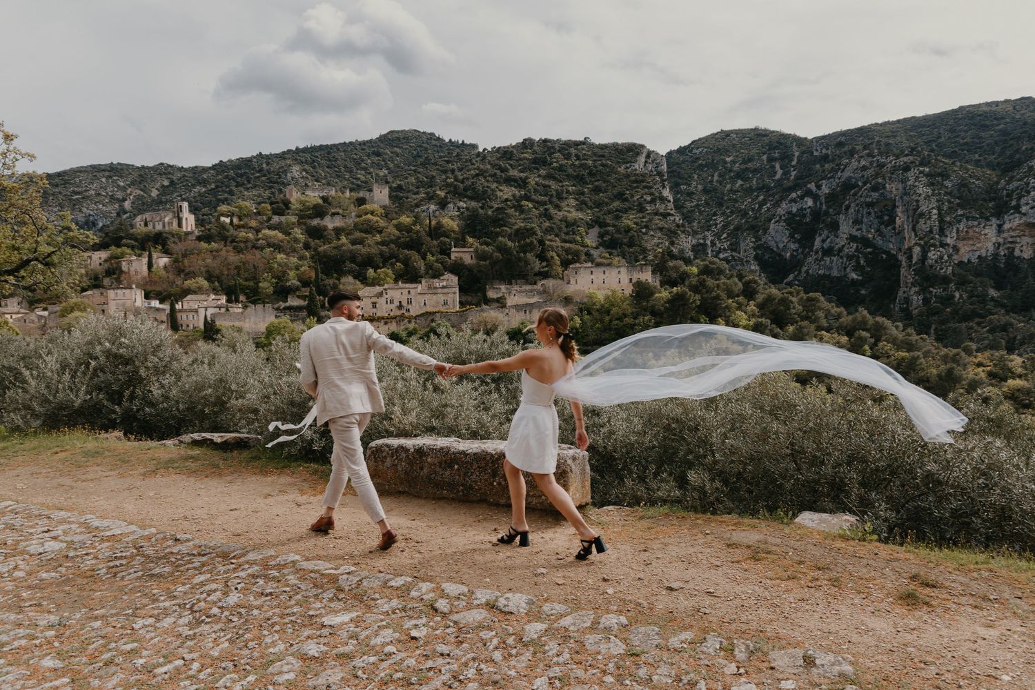 A couple in white wedding attire walks along a scenic mountain trail during sunset while their veil flows in the wind.