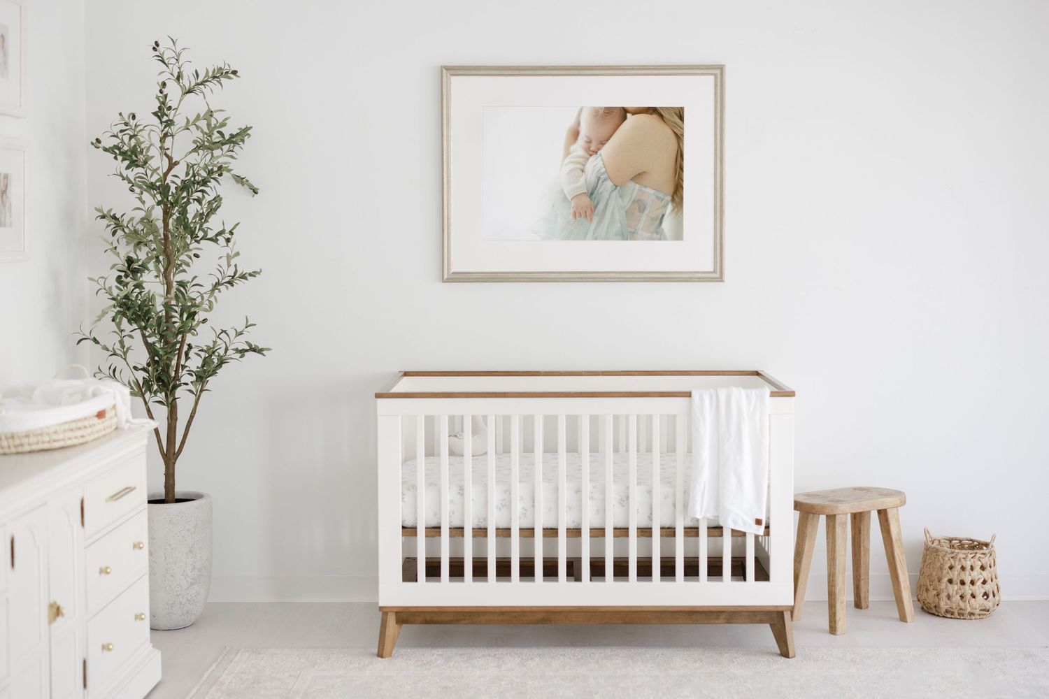 Minimalist Scandinavian style nursery with white crib, wooden furniture, and green plants against a white wall.