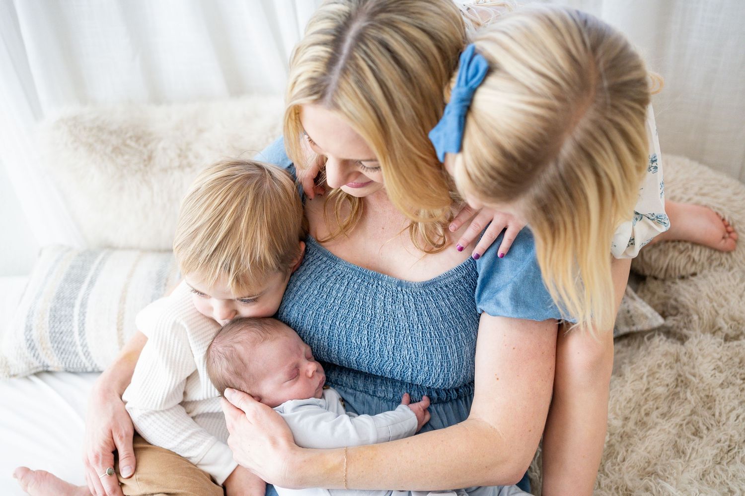 A tender family moment shows a mother in blue embracing two children on a cozy white blanket.