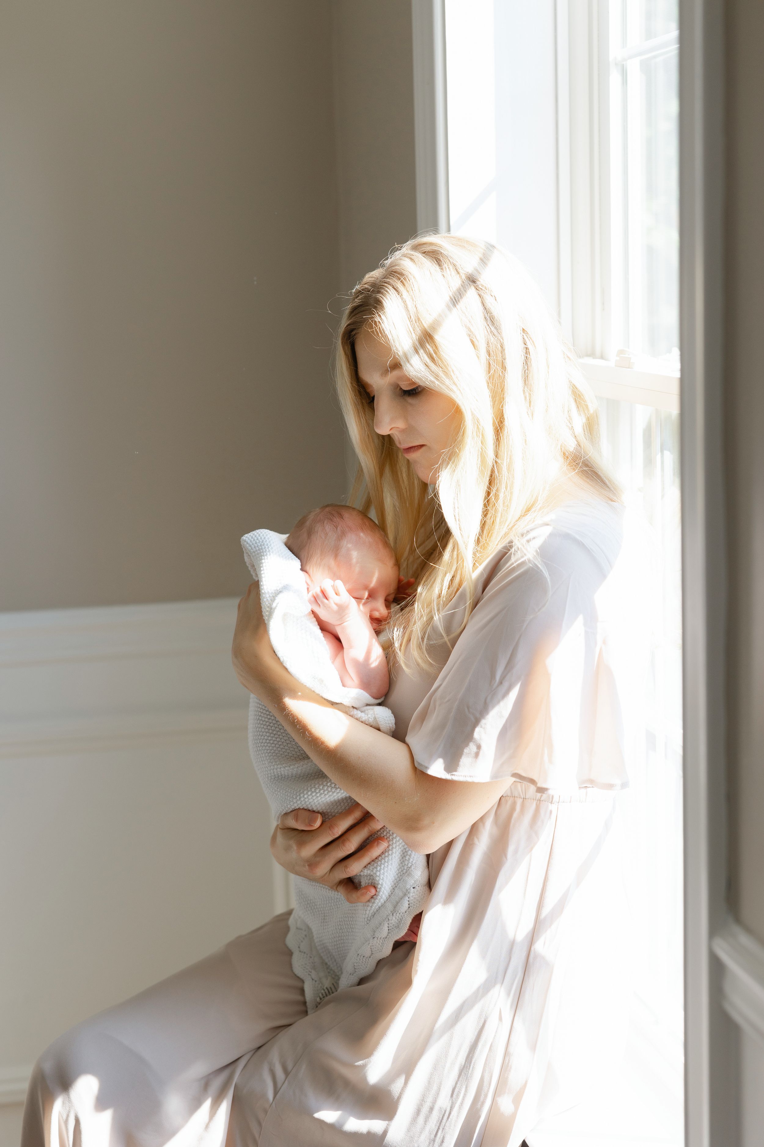 A tender moment between parent and baby in white clothing by a bright window.