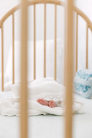 Tiny baby feet peek out from under a white knit blanket in a wooden crib.