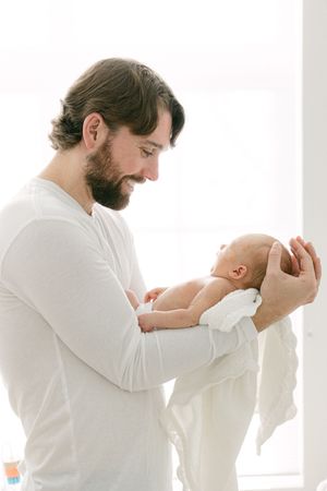 A tender moment between father and newborn baby in white clothing by a bright window.