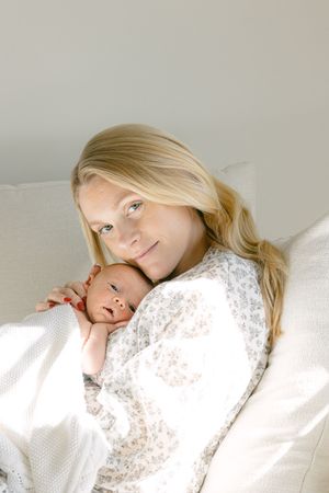 A mother in white lace rests with her newborn baby on soft white pillows.