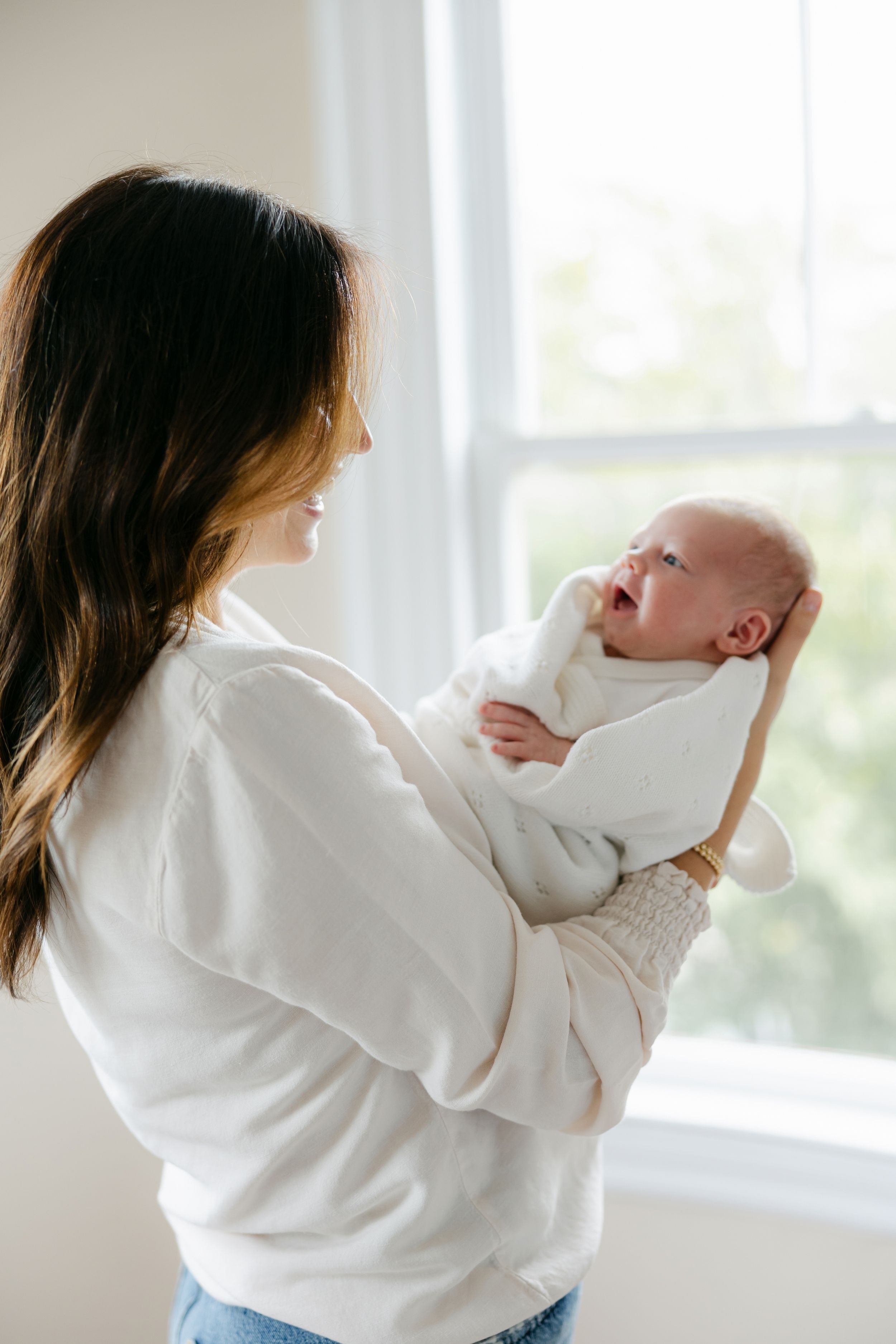 A parent in a white sweater holds a swaddled newborn baby next to a bright window.