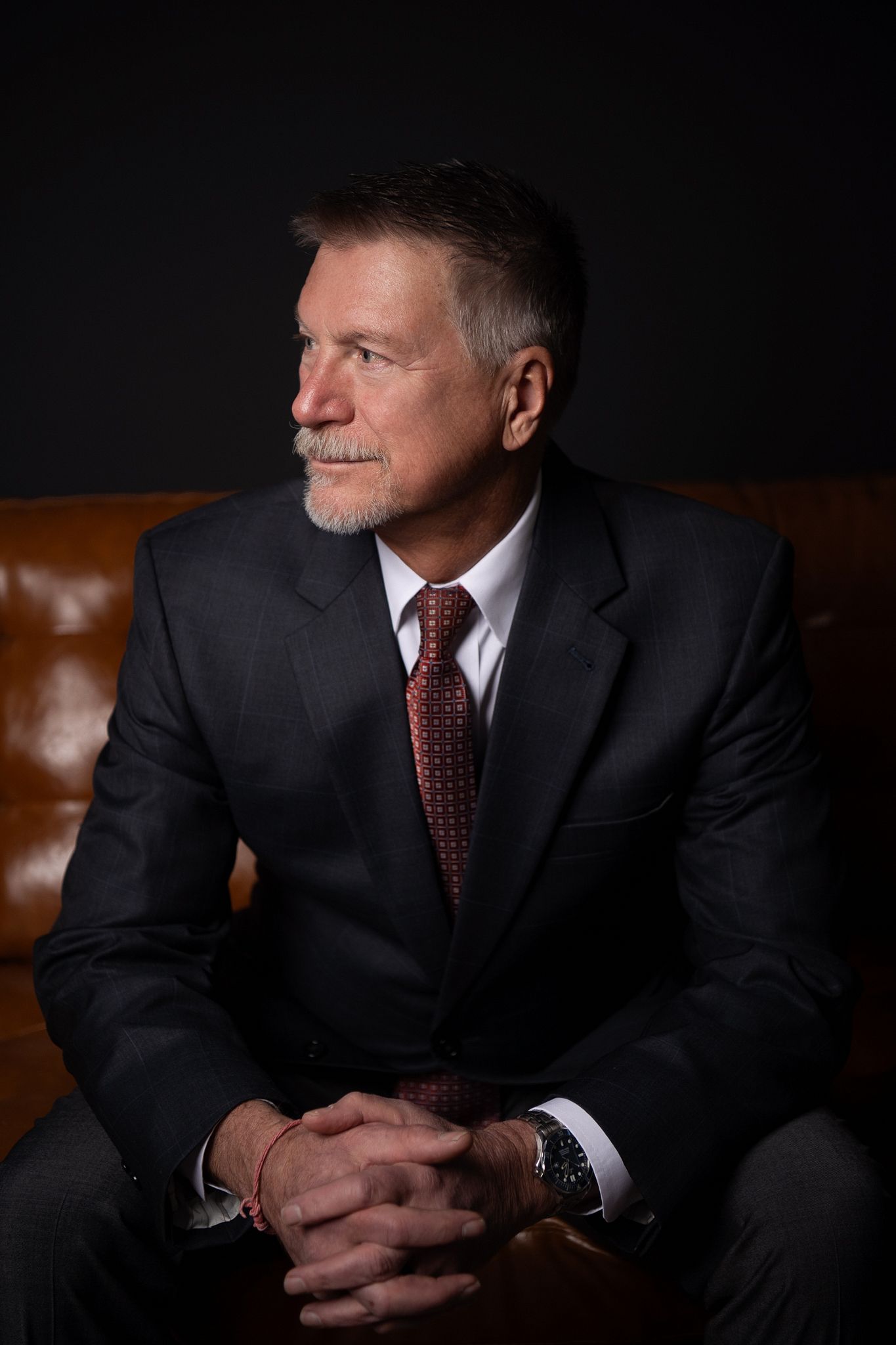 Professional business portrait series showing a person in dark suit and red tie seated on brown leather against black background.