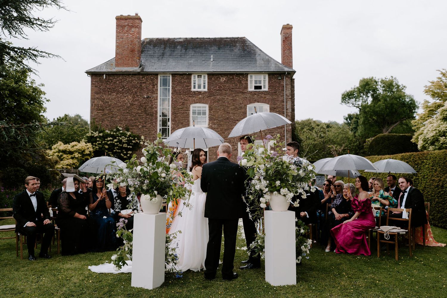 wedding guests, bride and groom sheltering underneath umbrellas during a rain shower during the ceremony at dewsall court in herefordshire