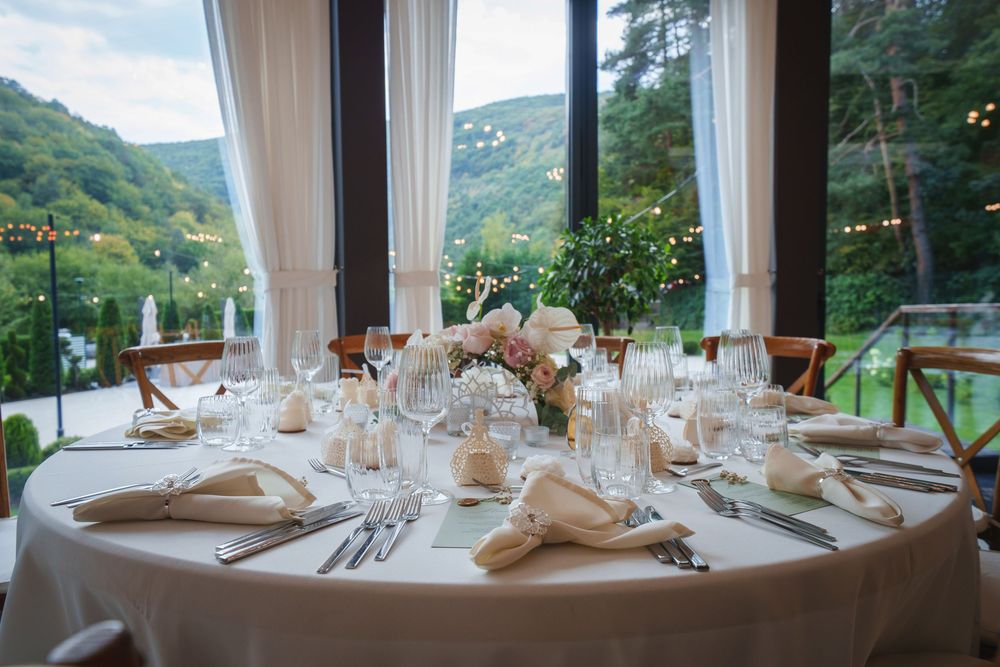 Elegant round dining tables set for a formal event with white linens and mountain views through panoramic windows.