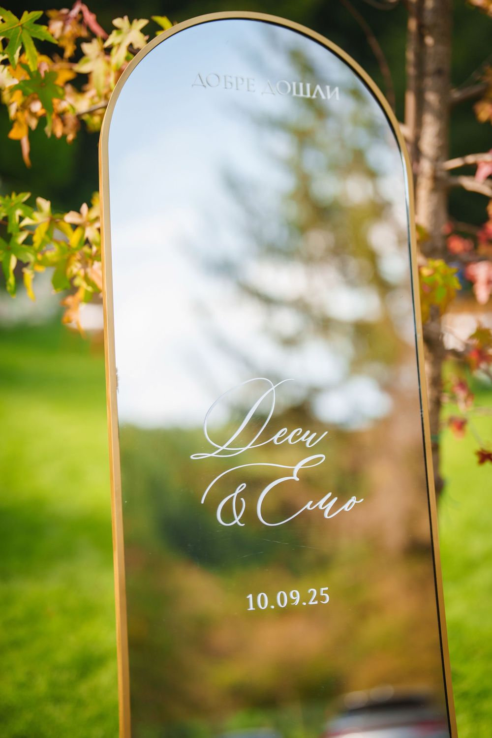 Elegant wedding mirror signage with calligraphy text displayed in an outdoor garden setting with soft bokeh background.