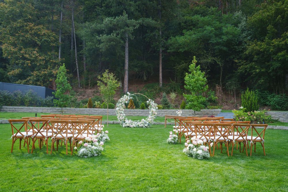 Rustic outdoor wedding ceremony setup with wooden chairs and floral arch on a lush green lawn surrounded by forest.