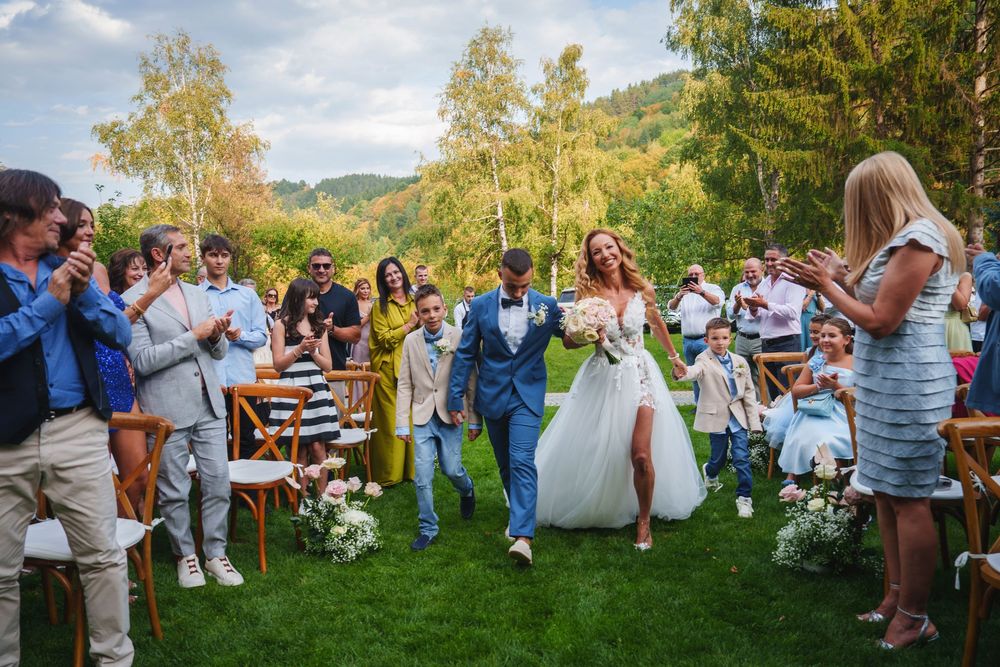 Wedding guests dance and celebrate outdoors at a rustic summer reception under blue skies and trees.