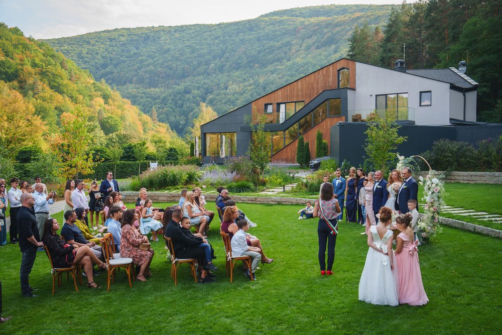 Guests gather for an outdoor wedding celebration on a lush hillside with mountains and autumn foliage in the background.
