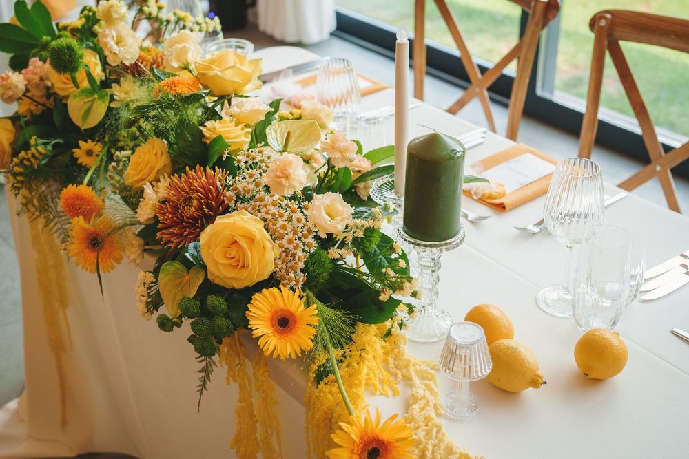 Elegant table setting with yellow and orange floral centerpiece featuring gerbera daisies and roses next to green candle.